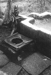 A pit laterine with a wooden toilet seat built next to a cement-covered mud-brick water trough.