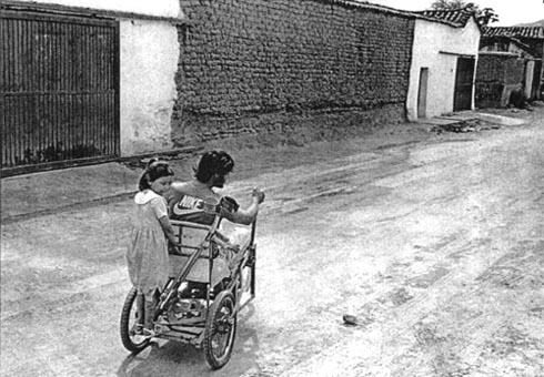A girl is riding on the back of the wheelchair driven by a man.