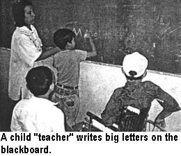 A child "teacher" writes big letters on the blackboard.
