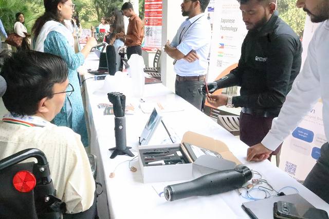 福祉機器企業の展示ブースの様子。テーブルには義手やIT機器が並べられ、担当者が来場者に義手のデモンストレーションを行っている。