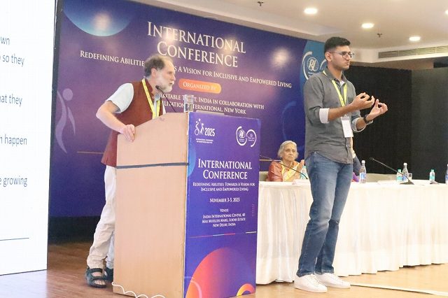 演台でスピーチをするイギリスからの講演者（左）と、その横に立ち手話通訳を行っている男性（右）。