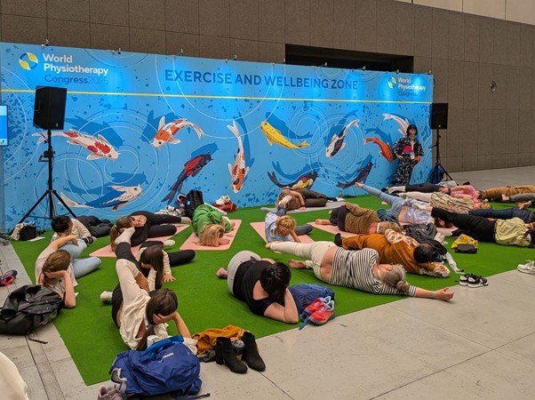 世界理学療法連盟学会2025（WPC2025）の「EXERCISE AND WELLBEING ZONE」で行われたヘルシーリビング・プログラムの様子。鯉が描かれた青いバックパネルの前で、参加者たちが芝生のようなマットの上で横になり、エクササイズを行っている。