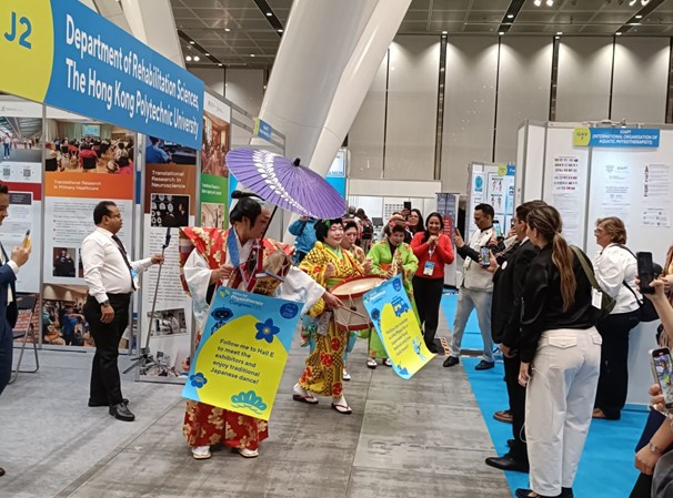 東京国際フォーラムの学会展示エリアで行われた、日本の伝統文化発信の様子。着物姿に和傘や太鼓を持ったチンドン屋が練り歩き、「Follow me to Hall E... enjoy traditional Japanese dance!」と書かれた案内板を掲げ、海外からの参加者を楽しませながら文化交流を行っている。