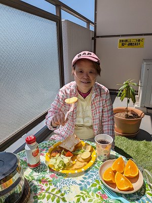 晴れた日のベランダで、パンやサラダ、オレンジなどが並んだランチを楽しんでいる真理さん