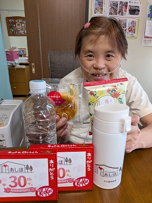「あしほ」30周年パーティーでもらった、記念の白い水筒やキットカットなどの前で微笑む真理さん